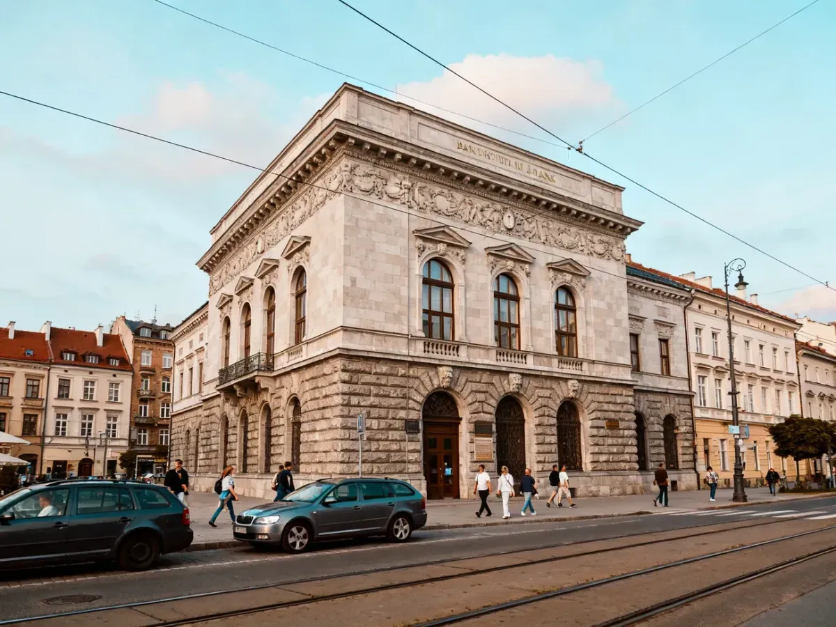 Zabytkowy gmach Banku Pekao w Krakowie z bogato zdobioną fasadą, ulicą z tramwajem, samochodami i przechodniami.