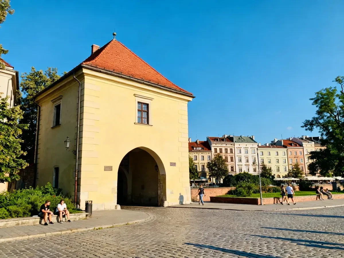Rogatka Pod Kasarnią w Krakowie w słoneczny dzień, żółta fasada z czerwonym dachem, brukowana ulica i spacerujący ludzie