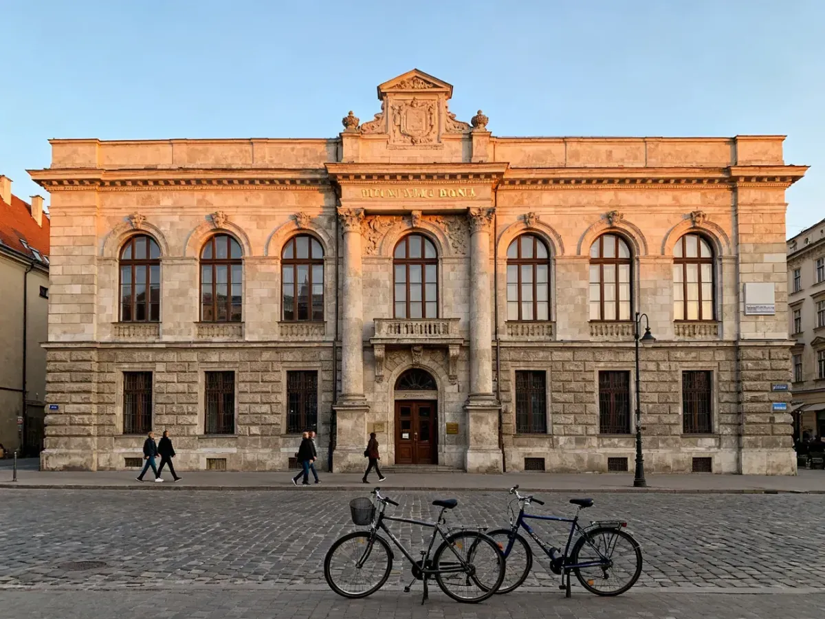 Historyczny gmach banku PKO w Krakowie w ciepłym popołudniowym świetle, z przechodniami i rowerami na brukowanej ulicy